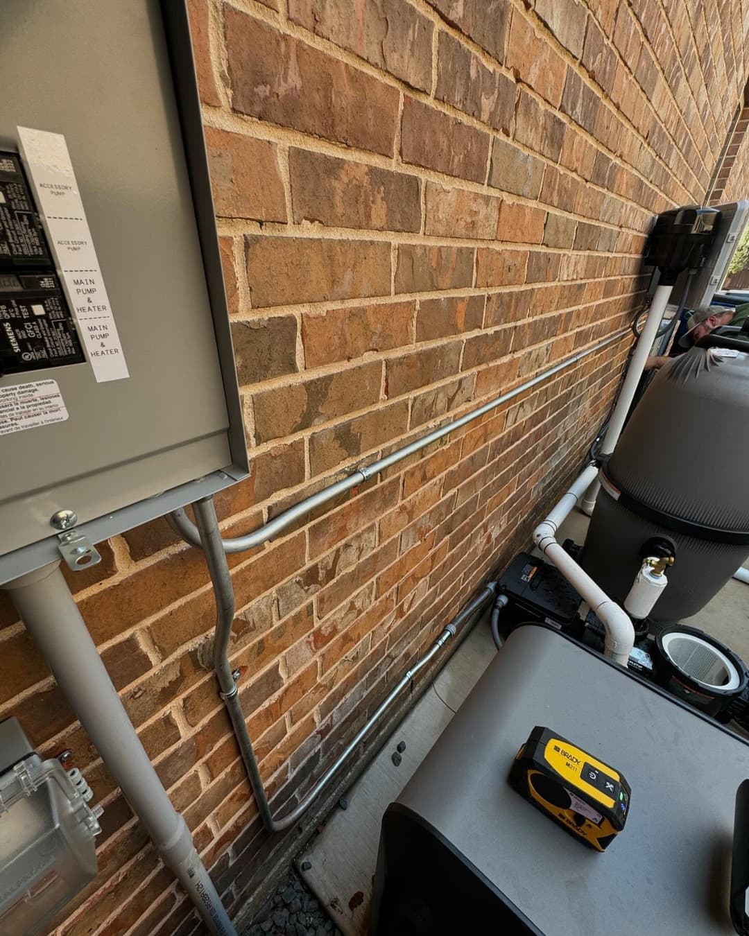 Electrical panel and plumbing pipes along a brick wall with tools on the ground.