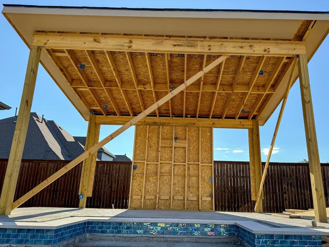 Open-air construction of a wooden pavilion with exposed beams and framing, near a pool.
