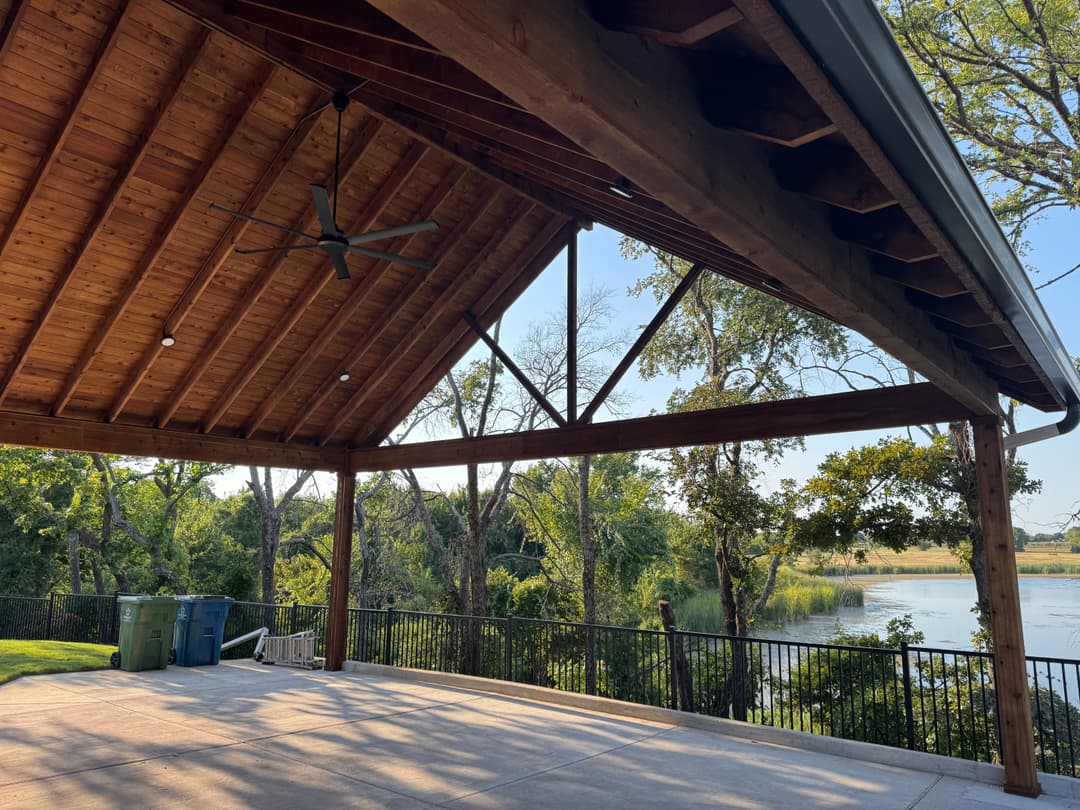 Wooden pavilion overlooking a serene lake, featuring a vaulted ceiling and ceiling fan.