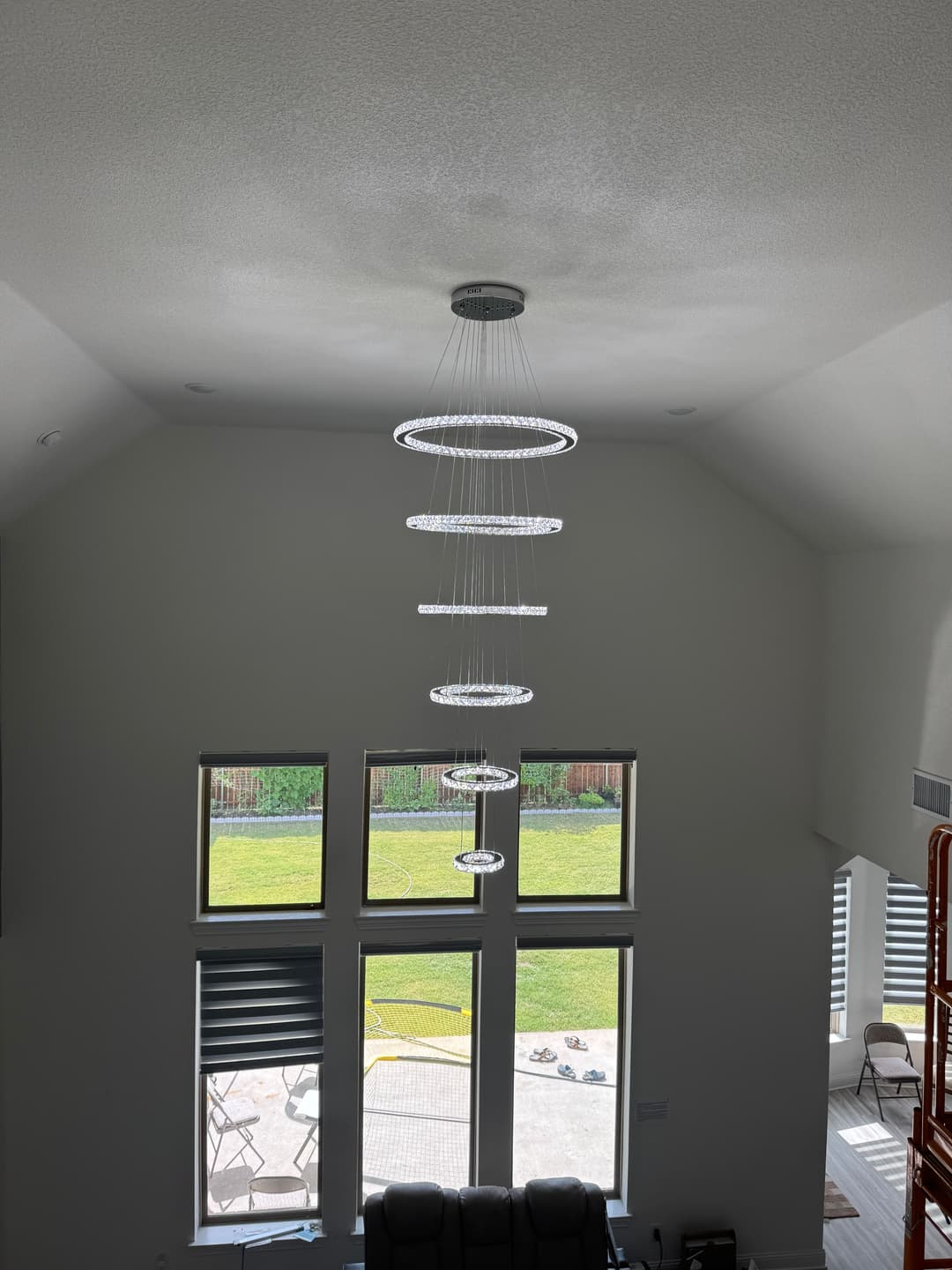 Modern chandelier hanging from a high ceiling in a spacious living room with large windows.