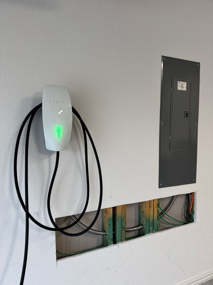 Tesla wall charger mounted on wall with electrical panel and exposed wiring.