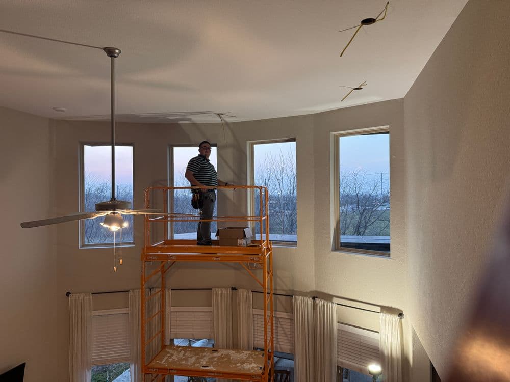 Worker on scaffolding installing fixtures in a room with large windows and modern decor.