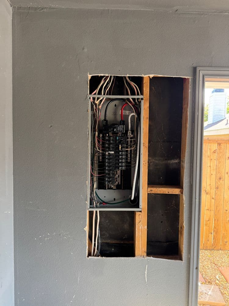 Exposed electrical panel in a wall cavity with visible wiring and gray interior paint.