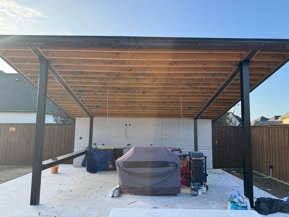 Modern outdoor patio with wooden roof, grill area, and construction materials visible.
