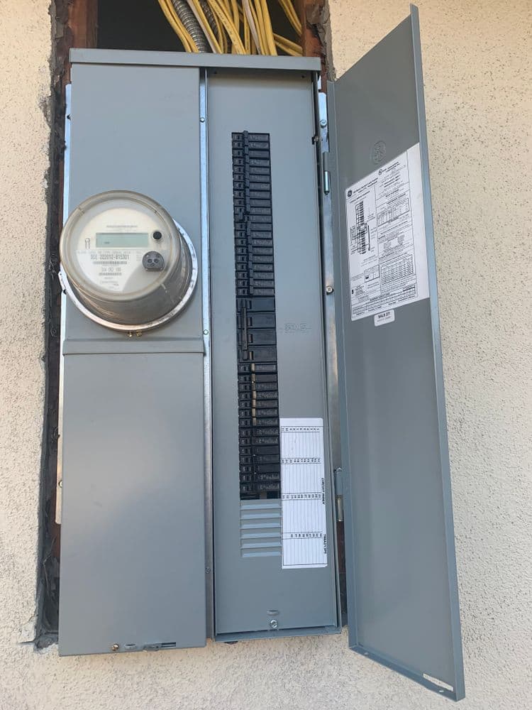 Electric meter and circuit breaker panel mounted on a wall, showing wiring and labels.