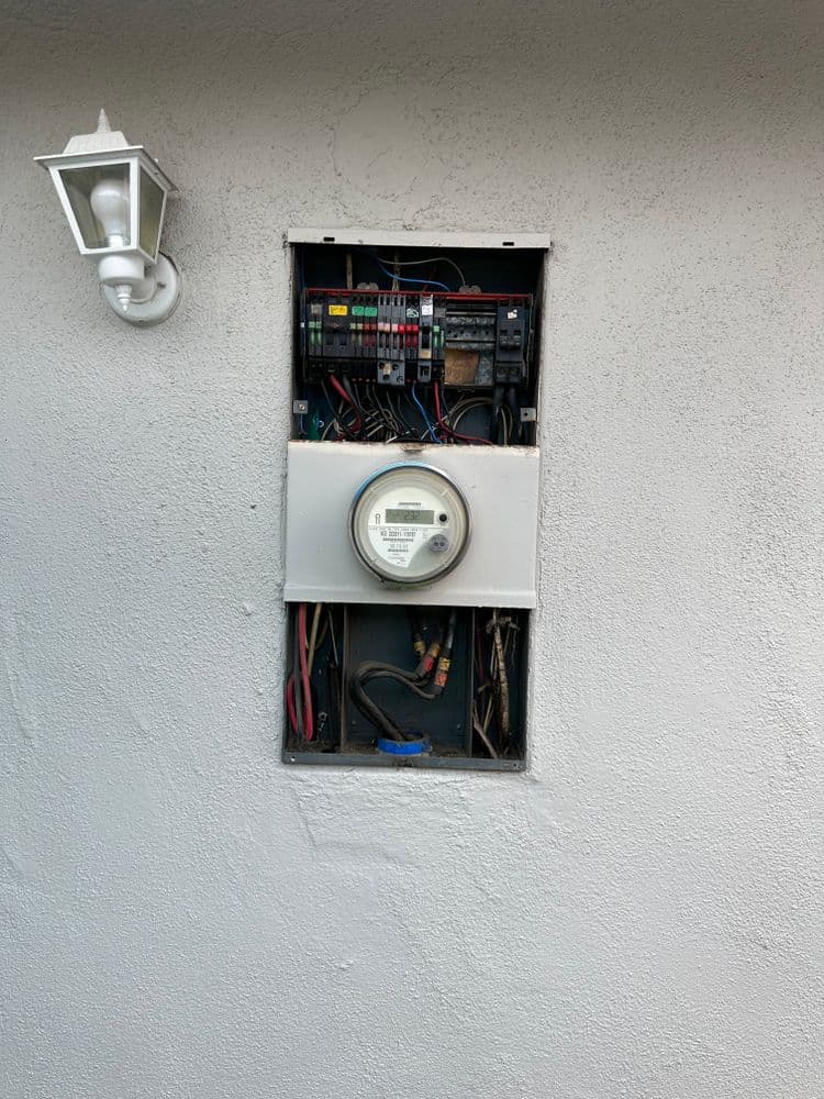 Electric meter and circuit breaker panel mounted on a textured white wall with outdoor light.