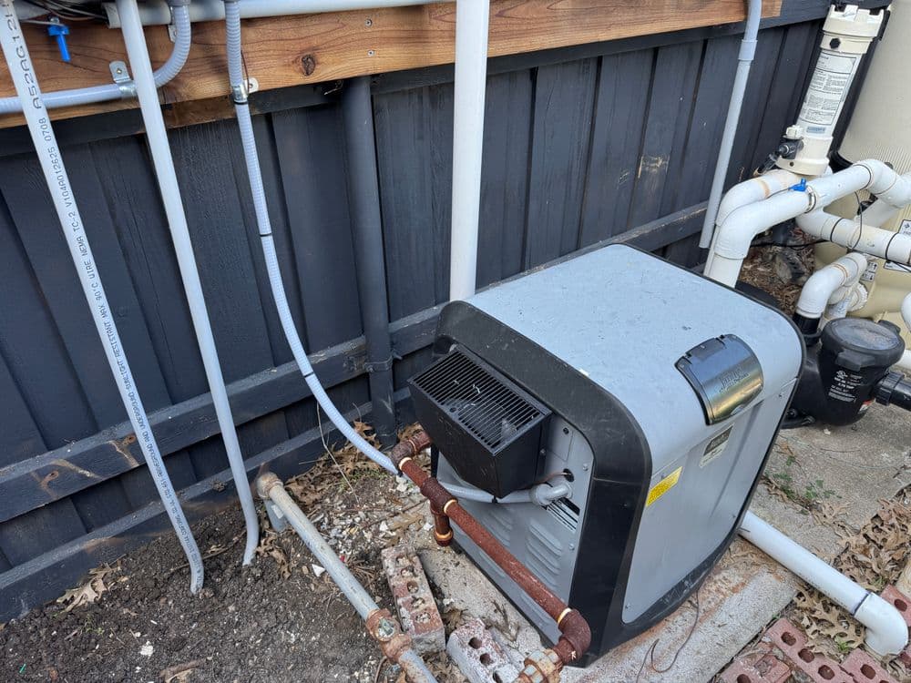 Outdoor gas pool heater installation with plumbing connections in a backyard setting.