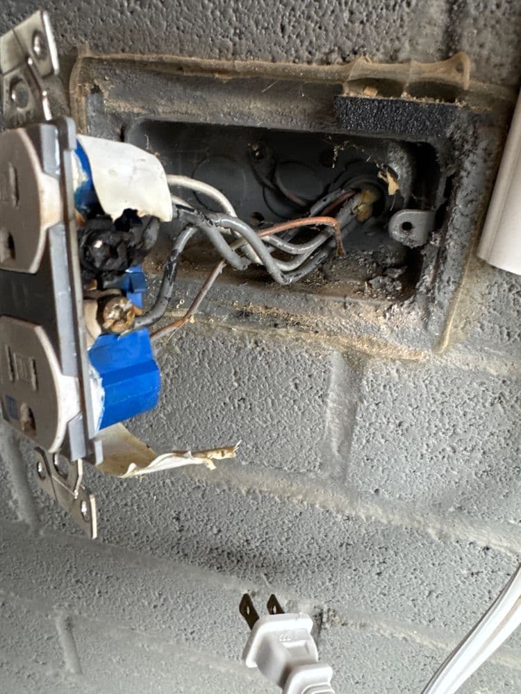 Damaged electrical outlet with exposed wires and burnt components on a gray wall.