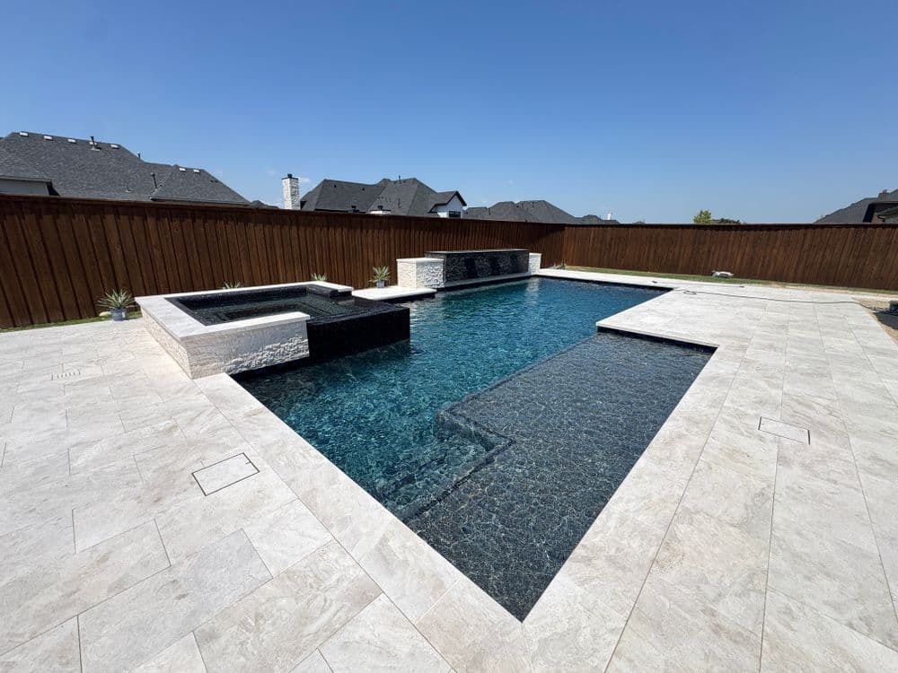 Luxury outdoor pool with spa, stone features, and wooden fence in a sunny residential backyard.
