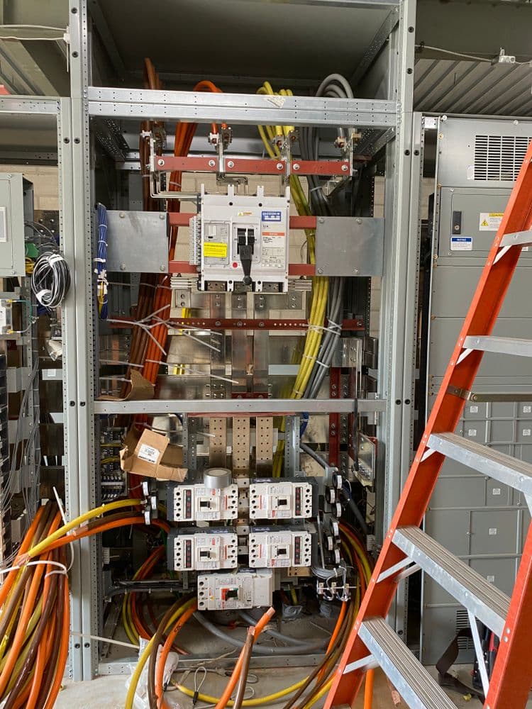 Electrical panel with circuit breakers, wires, and a ladder in a utility setting.