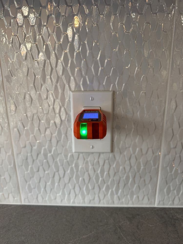 Smart energy monitor plugged into a wall outlet with a green status light. Honeycomb tile background.