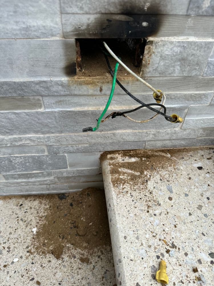 Damaged electrical wiring with burn marks near a stone wall and debris on the floor.